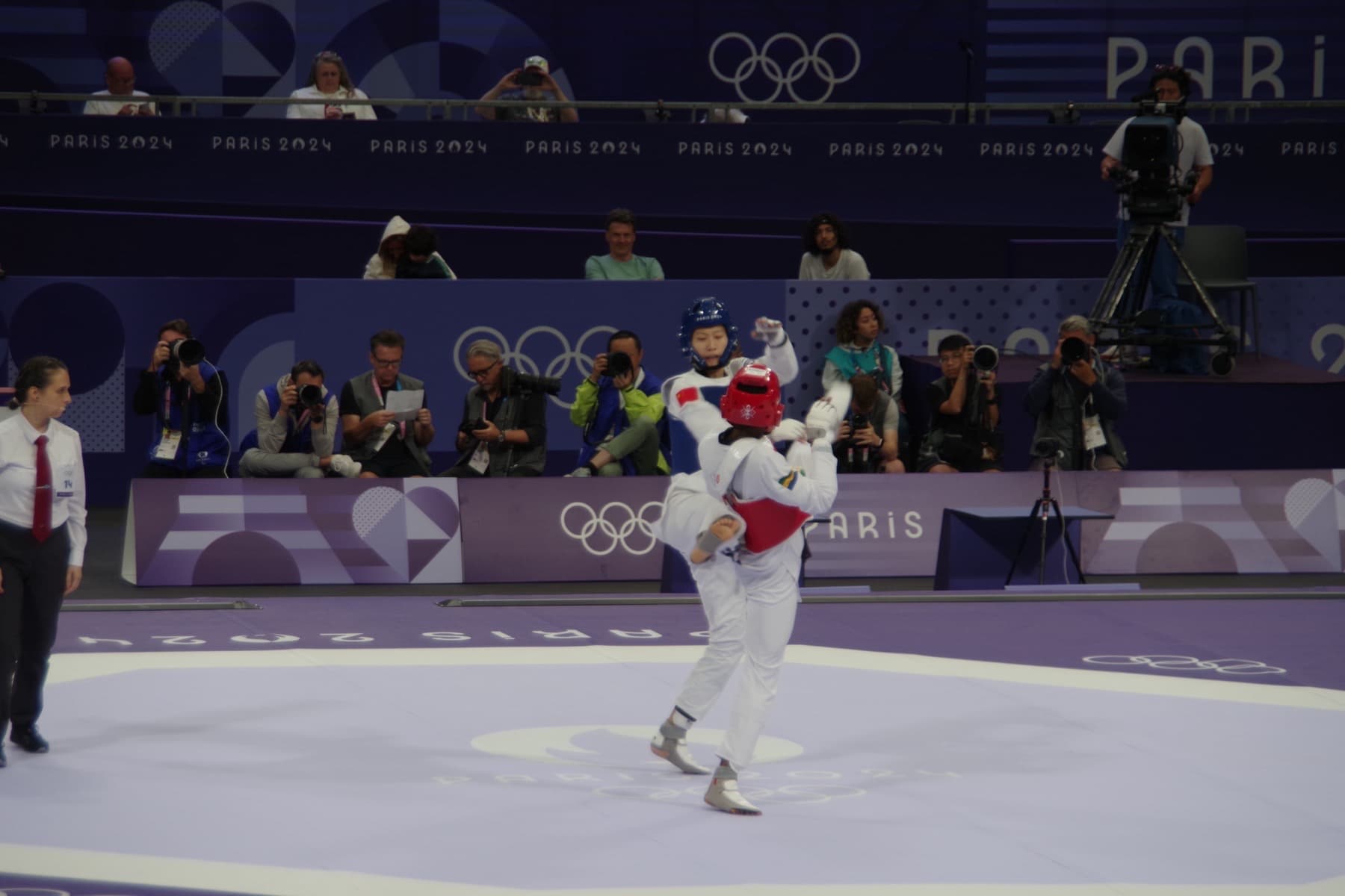 Women’s 57 kg taekwondo at the Paris 2024 Olympics in the Grand Palais — white dobok, electronic chest and head protectors, and the high kicks Virtual Taekwondo now tracks with motion sensors instead of impact scoring.