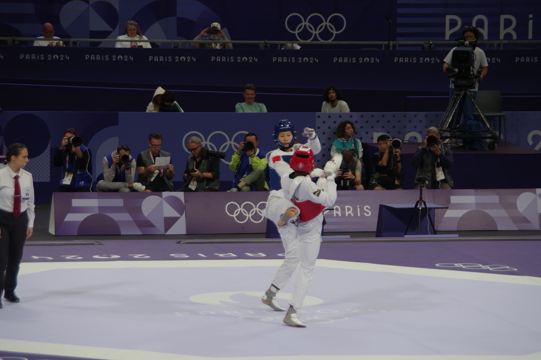 Women’s 57 kg taekwondo at the Paris 2024 Olympics in the Grand Palais — white dobok, electronic chest and head protectors, and the high kicks Virtual Taekwondo now tracks with motion sensors instead of impact scoring.